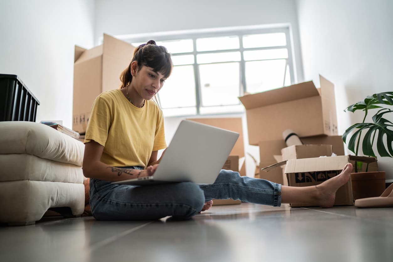 Young woman using laptop at new apartment, with moving boxes around.